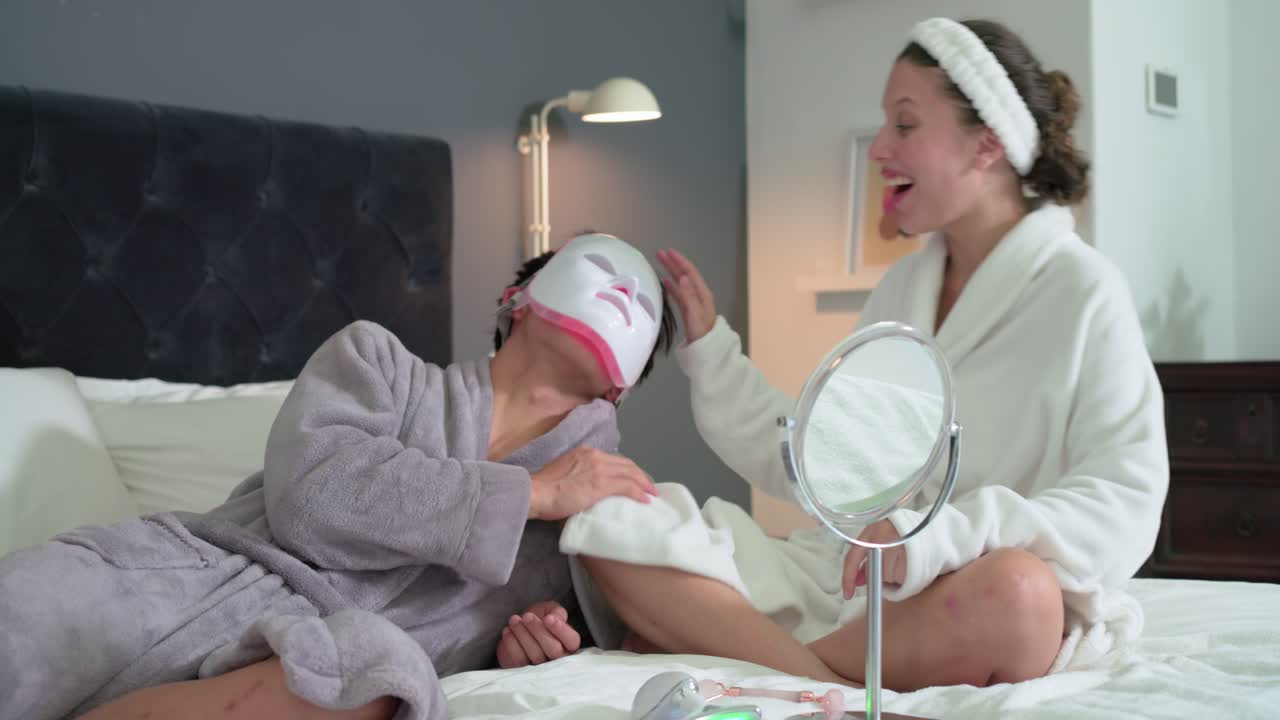 Diverse couple on bed while woman picking up, placing white LED mask, cycling lights for skincare