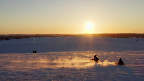 Snowmobiles-travelling-fast-across-snowy-sunlit-Swedish-Lapland-landscape-with-sunset-glowing-on-horizon