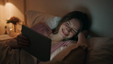 girl watching tablet online at night closeup. smiling woman resting bed enjoying