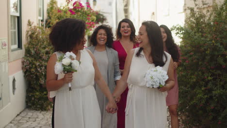 lesbian brides in white dresses with bouquets holding hands, laughing, talking with bridesmaids and walking in the street