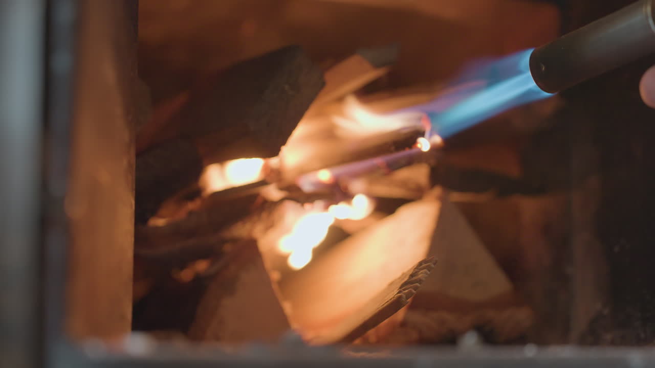 Close up inside chimney shows person igniting stacked fire wood with strong blue gas flame torch, kindling blaze among wooden logs, creating warm, glowing fire for indoor heat and comfort