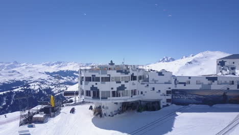 Pull-back-shot-of-men-enjoying-sunny-day-on-terrace-in-winter-mountain-centre.-Sporting-at-vacation-in-winter-landscape.-Laax,-Switzerland