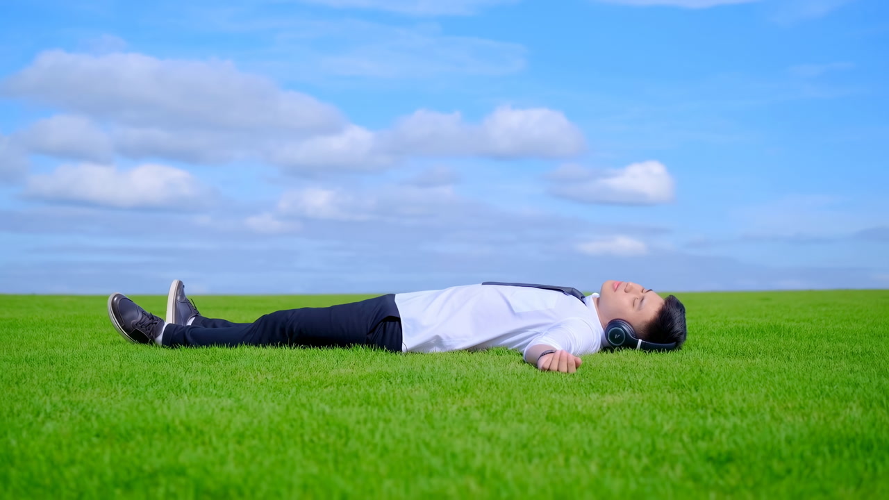 Man Lying On Green Grass Listening To Music Through Headphones, Wearing Suit And Tie, Eyes Closed Under Blue