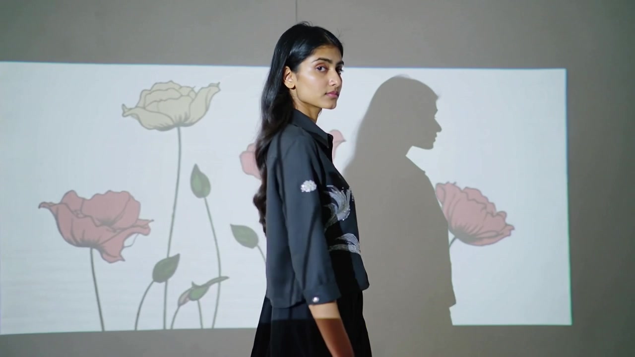 Una modella indiana cammina attraversando lo studio in giacca e gonna, con una proiezione floreale e un'ombra sovrapposte.