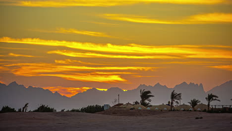 Orangefarbener-Himmel-Mit-Wolken-Bei-Sonnenuntergang-über-Einer-Wüstenlandschaft-Mit-Palmen-Und-Bergen-Am-Horizont