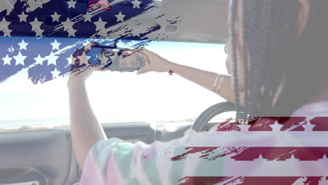 animation of flag of usa over african american woman in car by beach in summer