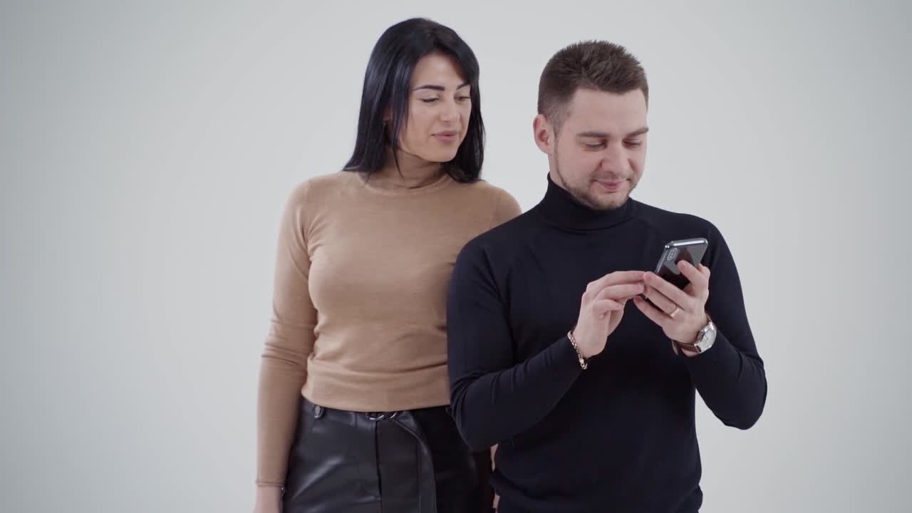 Handsome man looking at the phone in studio. Beautiful young woman standing behind the man. Young couple on the white background.