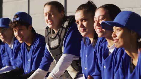 un grupo feliz y diverso de jugadoras de béisbol sentadas en el banco escuchando al entrenador y sonriendo