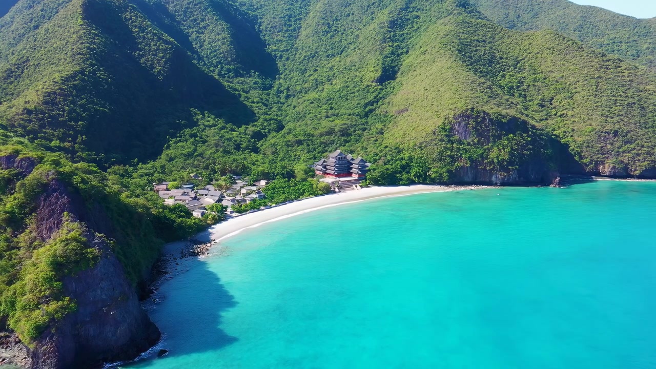 Vista aérea de una serena bahía tropical con agua turquesa brillante, una playa curva de arena blanca, montañas verdes escarpadas y un pequeño pueblo costero cerca de la orilla.