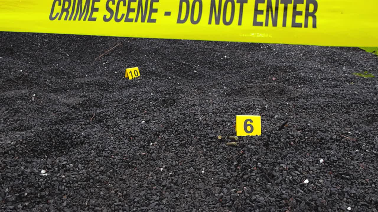 Yellow police line or crime scene forensic investigation tape in front of a dark gravel outdoors area where Crime scene markers are next to evidence like high caliber bullet casing and ammunition