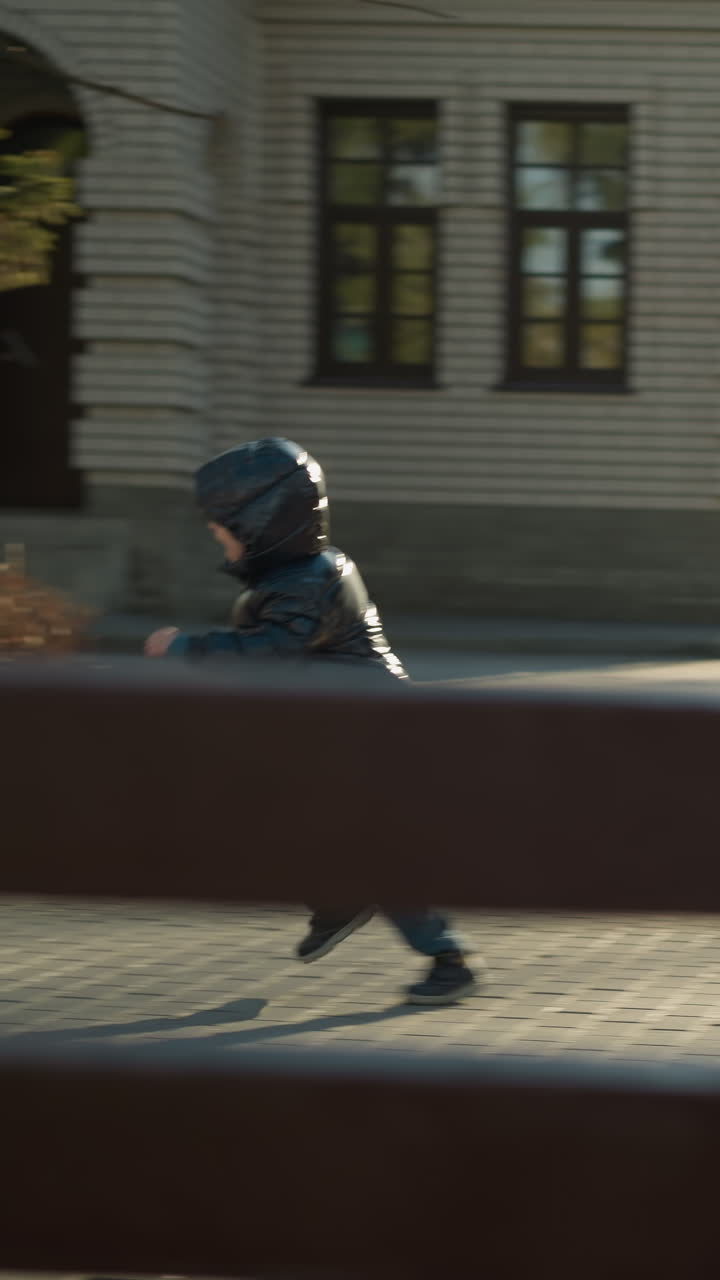 Father and son chasing a balloon along a paved path near a building, the child, dressed in a shiny black jacket, reaches out to catch the balloon while the father approaches with open arms