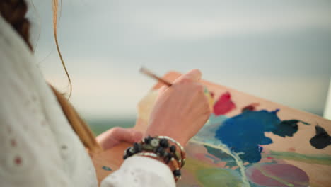 un primer plano de la mano de una mujer, adornada con cuentas, mezclando suavemente colores en una paleta con un pincel. la escena captura el delicado movimiento de mezcla de pintura bajo la suave luz del sol