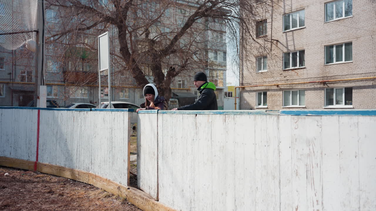 Два белых подростка на открытом катке смотрят через ограждение, многоквартирный дом на заднем плане, куртки с капюшоном, идут вдоль потертого белого барьера, несут небольшой футбольный мяч, непринужденная уличная атмосфера, приглушенная зима