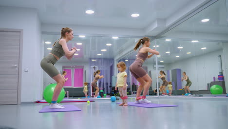 two women in a gym perform squats with resistance bands on their legs, stretching and toning muscles, a toddler, also with a band, watches them intently, surrounded by colorful gym equipment