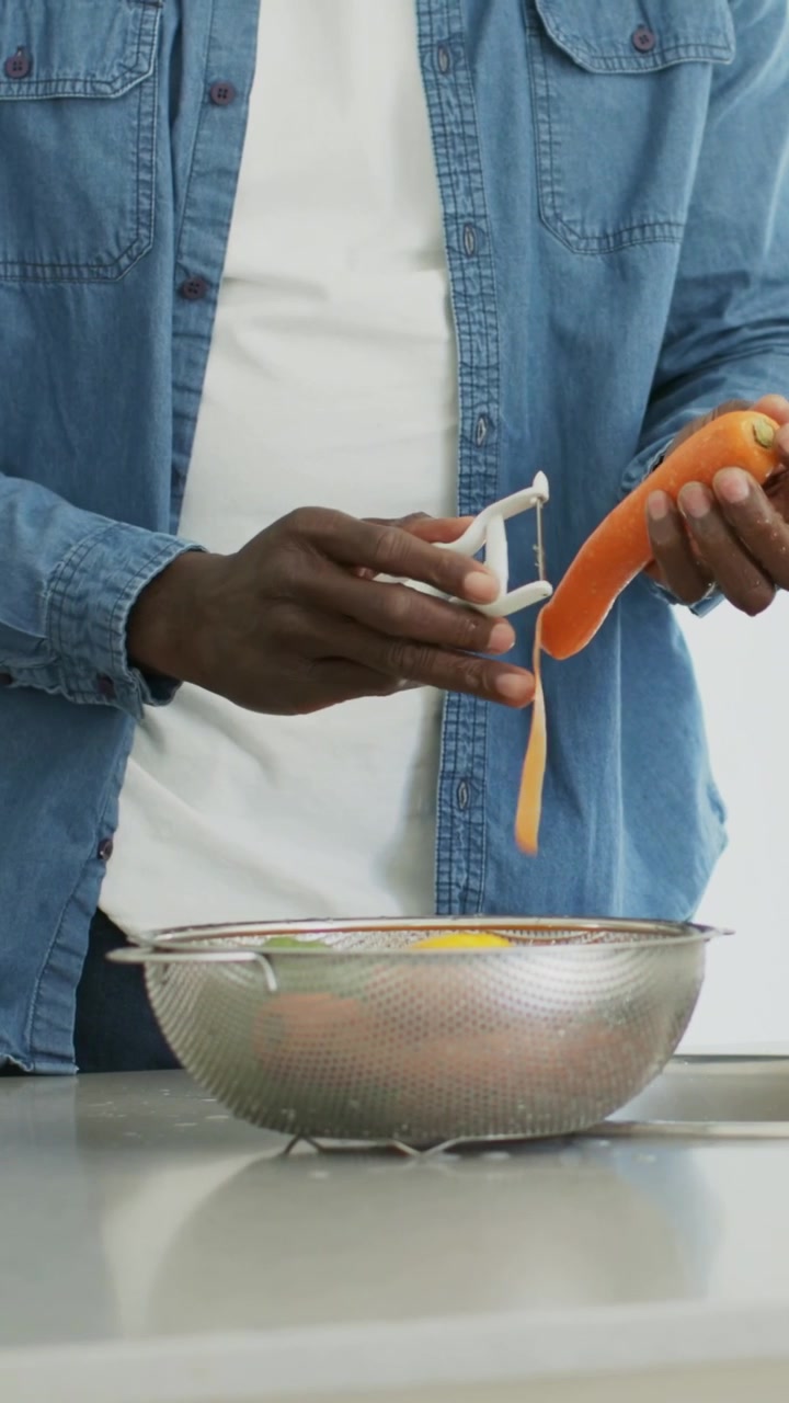 Vertikales Video: Mann im Jeanshemd schält eine Karotte für eine Mahlzeit an der Arbeitsplatte, daneben ein Sieb.