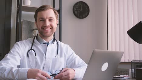 the family doctor sits in the clinic at the laptop, he takes off his glasses and laughs looking at the camera.