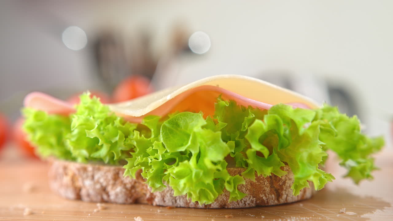 una rebanada de queso cayendo sobre un sándwich hecho con pan blanco, jamón y lechuga, cámara lenta y macro