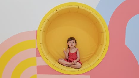 Cute-Kid-Girl-Eating-Lollipop-Sits-Cross-legged-in-Circular-Yellow-Wall-Built-in-Soft-Sofa