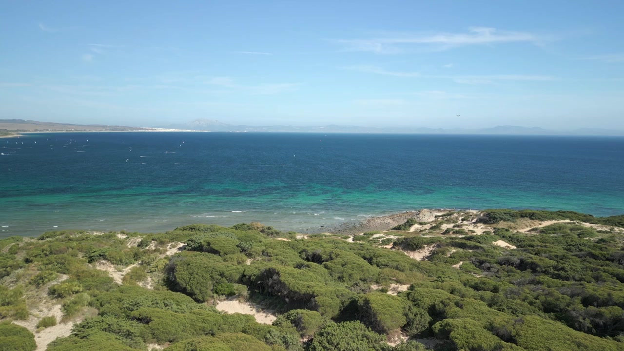 Sorvolo rapido e a bassa quota sulla vegetazione dunale costiera che si apre sulle acque dell'oceano piene di kitesurfer attivi a Tarifa in una giornata limpida. Ripreso in 4K a 25 fps