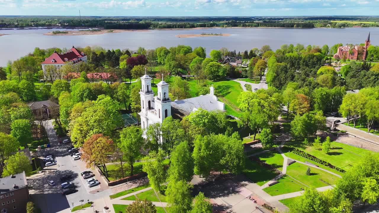 Drone footage of a white church with twin towers located in a lush green park within a scenic riverside town, with surrounding streets and distant islands under clear skies.