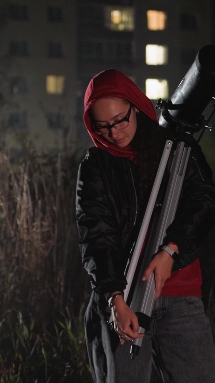 Giovane donna con cappuccio rosso regola il treppiede del telescopio all'aperto. La donna maneggia le parti ripiegate del telescopio. L'ambientazione include un primo piano erboso e finestre illuminate in lontananza