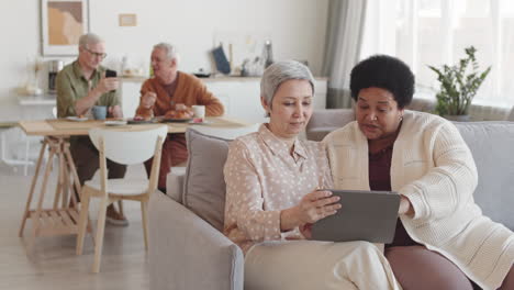 medium long of senior asian and african female friends sitting on couch in living room, watching something on tablet computer, smiling, talking in foreground of men communicating
