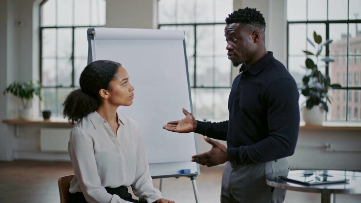 Starting standing man coaching seated woman in office, wearing business casual, flipchart