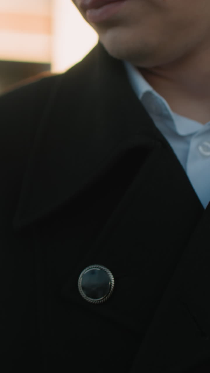 primer plano de un hombre sosteniendo un teléfono para responder una llamada, vestido con un traje negro con botones metálicos, camisa blanca debajo, ambientado contra un fondo borroso con cálidos reflejos de luz solar en un edificio de vidrio