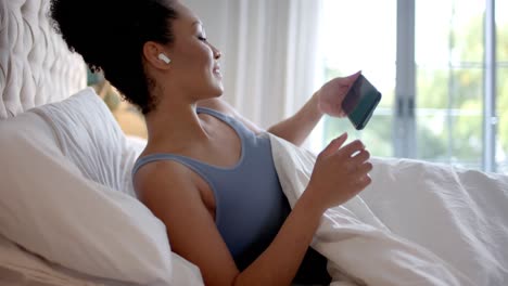 Biracial-woman-with-earphones-lying-in-bed-and-using-smartphone-in-bedroom,-slow-motion