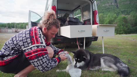 male camper pours dog food into a plastic bowl for his pet alaskan malamute