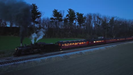 drone slightly ahead and parallel night view of a steam passenger train stopped in farmlands blowing lots of smoke seeing the lights in the coaches