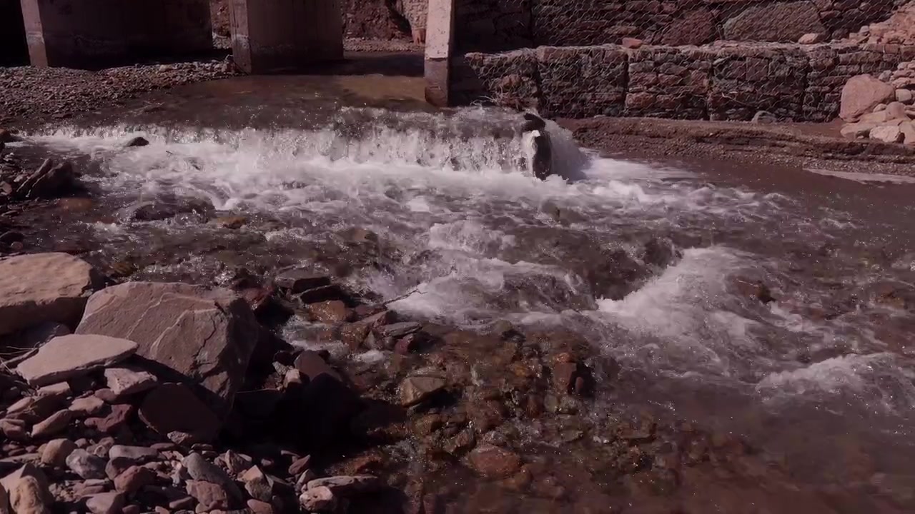 Corrente de rio caudalosa fluindo sobre um açude rochoso com um muro de pedra antigo e uma estrutura de concreto em Marrocos.