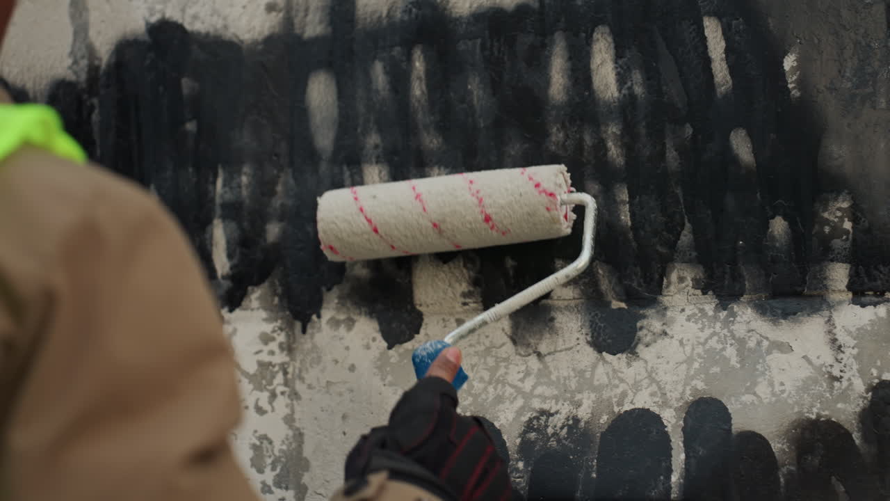 Primo piano di un rullo che copre graffiti neri, operaio sud-asiatico che ripara un muro di cemento, indossando un giubbotto ad alta visibilità e guanti, pennellate orizzontali metodiche su una superficie ruvida e incrinata, primer.