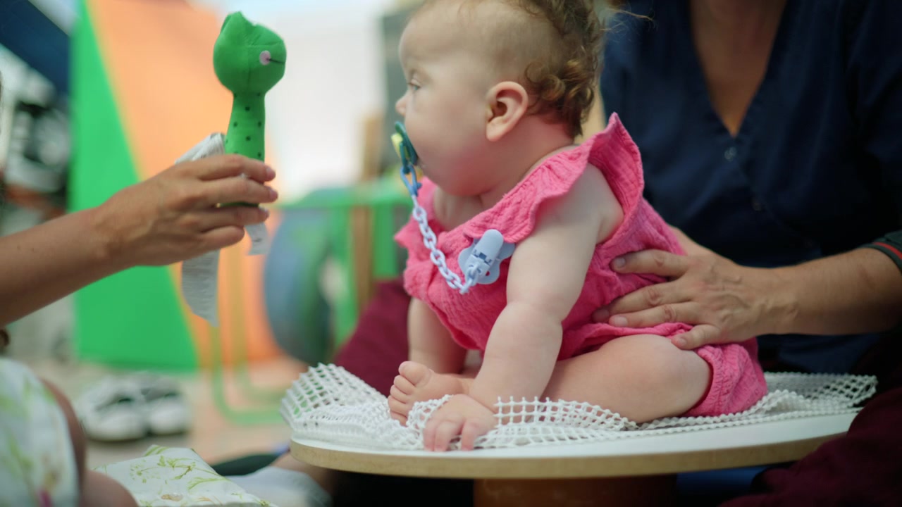 Bebê menina sentada em um suporte, recebendo intervenção precoce e fisioterapia com um terapeuta estimulando-a com um brinquedo.