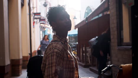 Close-up-of-a-young-black-woman-with-short-hair-standing-on-a-street-looking-up-and-turning-to-camera,-side-view,-backlit