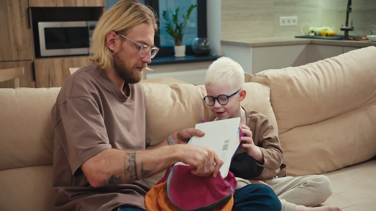 A blond man in glasses with a beard helps his little son, an albino boy with white hair color in blue glasses, collect his bright briefcase for school while preparing his homework at home in a modern apartment