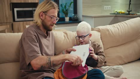 A-blond-man-in-glasses-with-a-beard-helps-his-little-son,-an-albino-boy-with-white-hair-color-in-blue-glasses,-collect-his-bright-briefcase-for-school-while-preparing-his-homework-at-home-in-a-modern-apartment