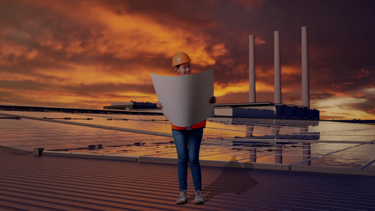 Helkropsbillede af asiatisk kvindelig ingeniør med sikkerhedshjelm, der kigger på en tegning i sine hænder, mens hun står og inspicerer solpaneler på et tag ved solnedgang.