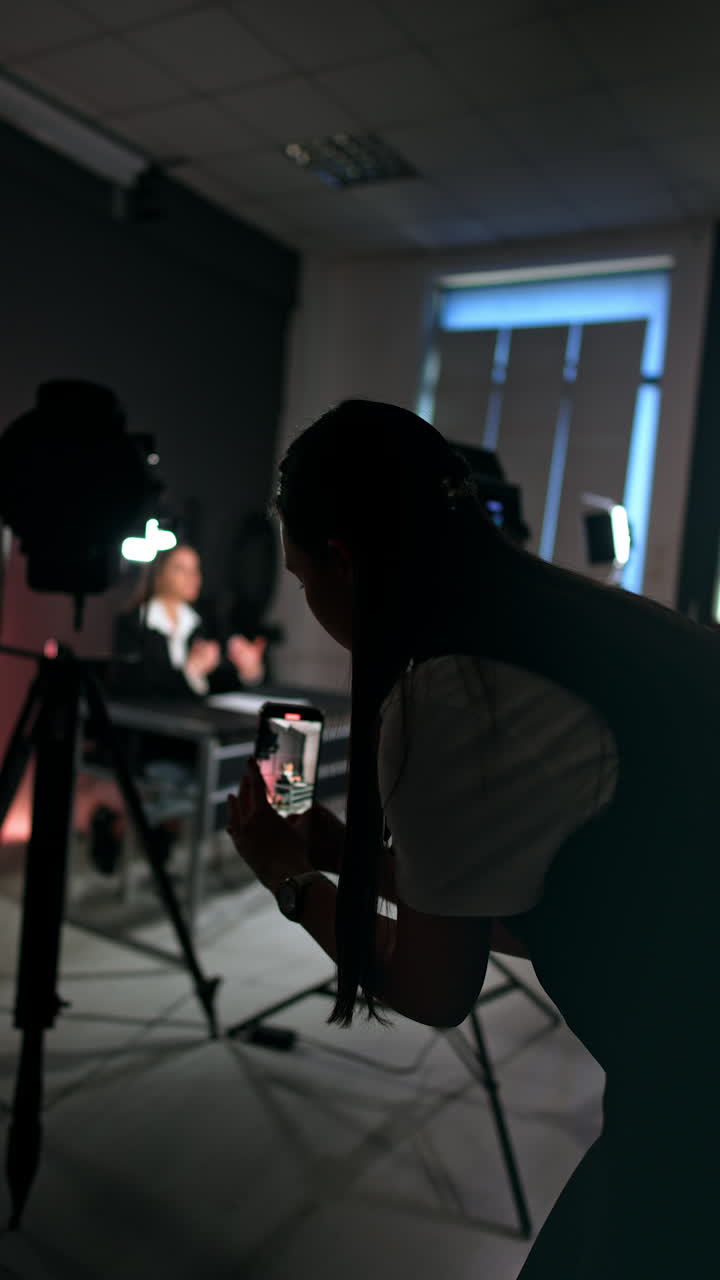 Long-haired woman taking video on her phone. Female reporter sitting at desk talks to camera. Vertical video.