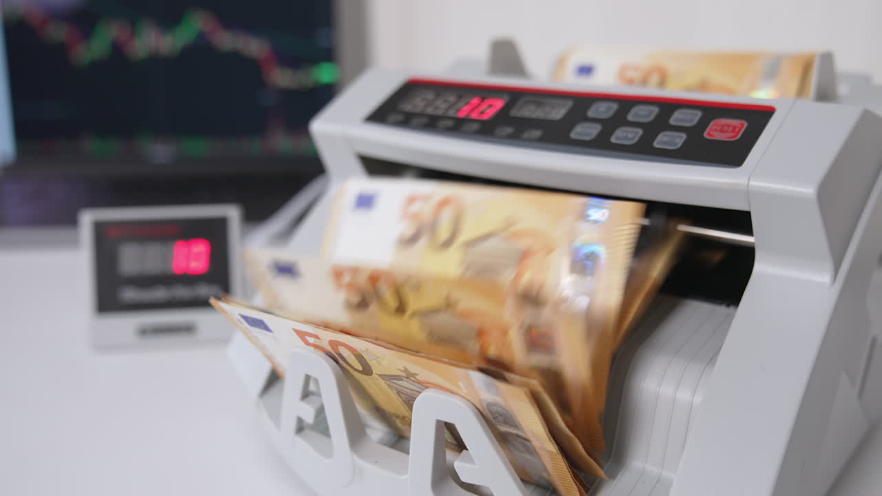 Hand places a wad if fifty euro bills into a slot of cash counting machine. Counter and switched on laptop at backdrop in blur.