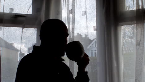 silhouette of bald male standing beside the window, as he savors his coffee while gazing at the daytime scene outside, exemplifying the essence of routine household moments