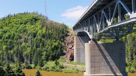 a drone flies across a river with a large bridge on the right side surrounded by trees, forests and mountains