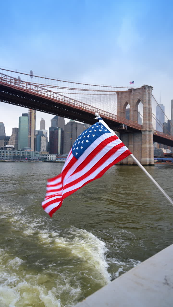 Bandiera degli Stati Uniti su traghetto con lo sfondo del Ponte di Brooklyn. Bandiera americana sventolante sul ponte di una barca che passa davanti a uno storico ponte in pietra e ai grattacieli di Manhattan