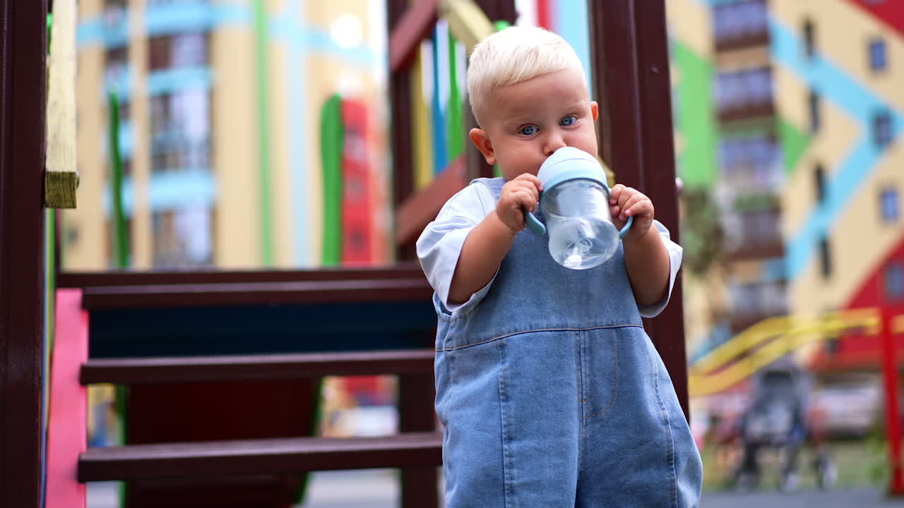 Lovely toddler boy in a loose jeans romper is at the playground. Kid picks up a water bottle and drinks from a straw. Blurred backdrop.