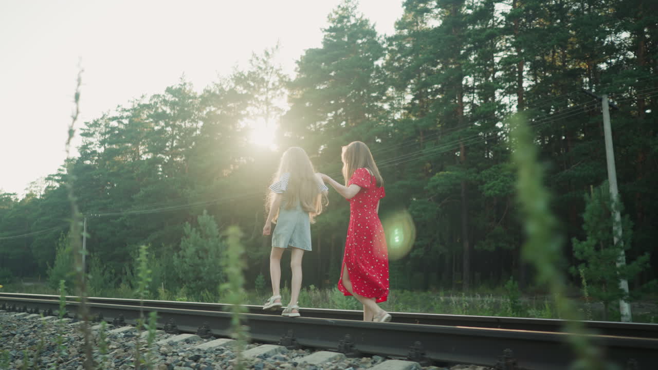 back view of woman in red dress holding little girl hand while she walks on rail beam during bright sunny day, with sunlight flare creating dreamy effect and power lines