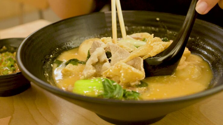 Una persona mescola una zuppa di noodle piccante con maiale, verdure, bacchette e cucchiaio in un ambiente con illuminazione calda