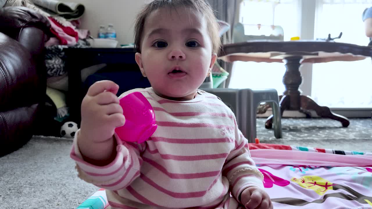 A curious baby sits on a rug indoors, holding a bright pink cup in a cozy home setting