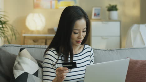 Mujer-Joven-Con-Una-Blusa-A-Rayas-Con-Una-Laptop-En-El-Regazo-Y-Compras-En-Línea-Con-Tarjeta-De-Crédito-Sentada-En-Un-Sofá-En-La-Sala-De-Estar-1