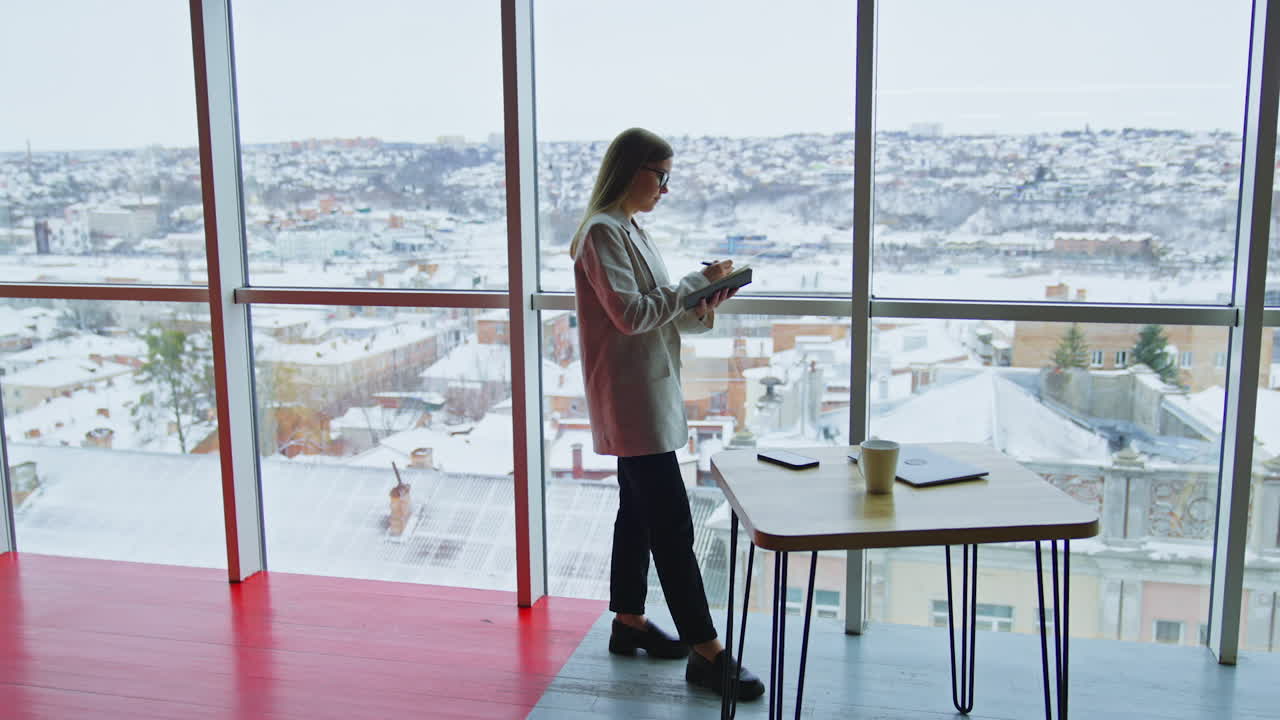 Happy smiling woman taking notes into a notebook. Lady standing near the table in front of panoramic windows with cityscape.