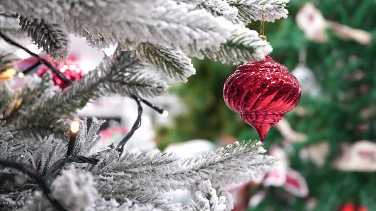 Close up slow motion shot of a red glass ornament swaying gently on a snow-dusted artificial tree branch with lights